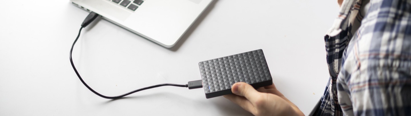 A hand connecting a portable hard drive to a laptop on a white desk.